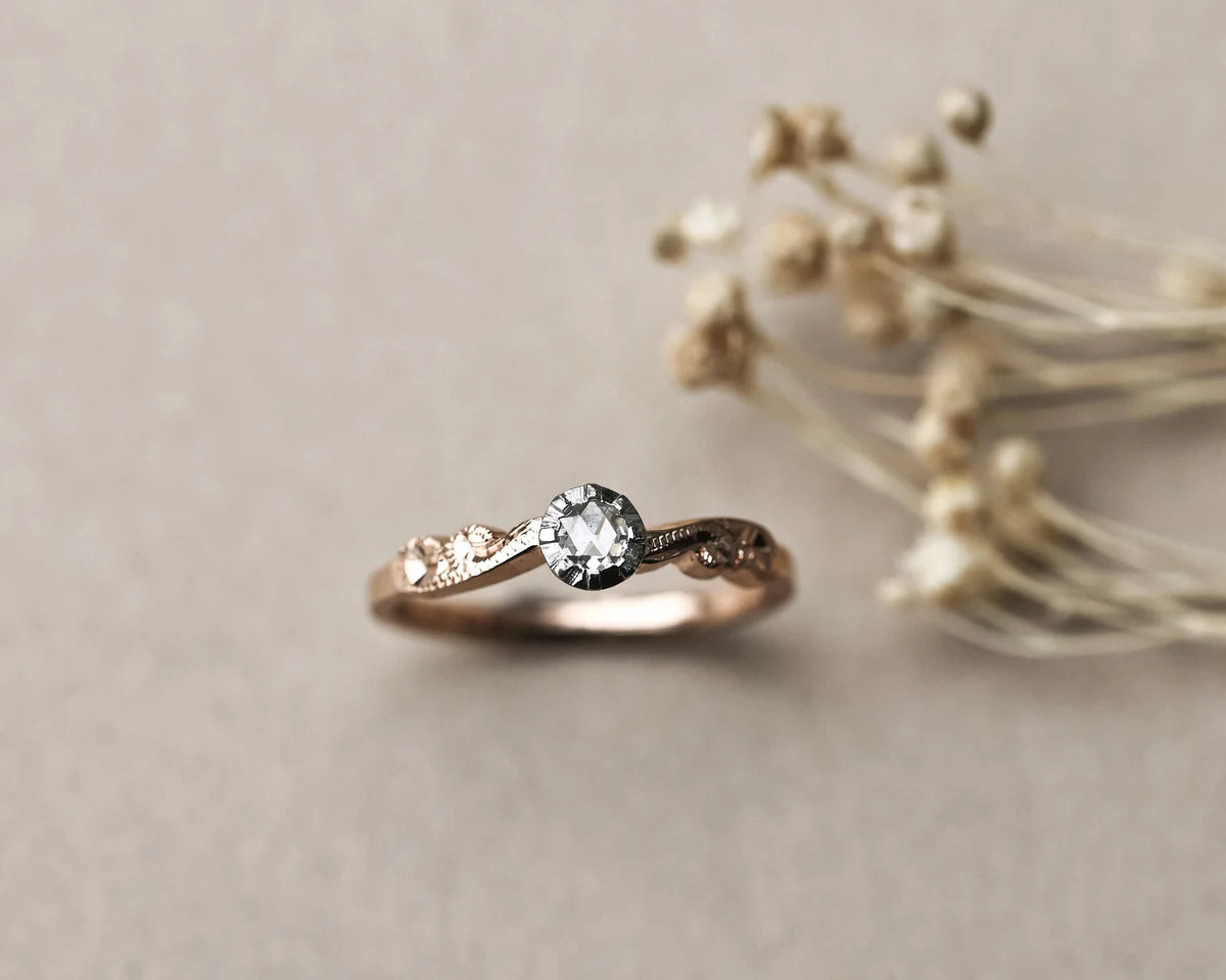 A rose gold engagement ring with a central round-cut clear gemstone and small side stones, styled on a neutral beige background with dried flowers.