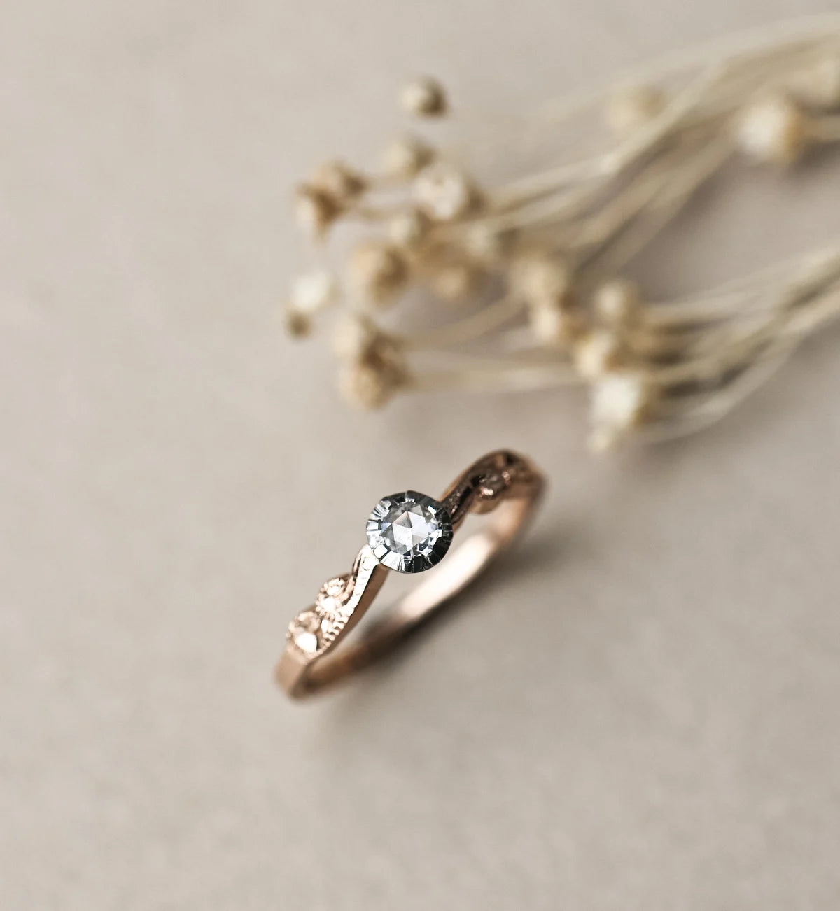 A delicate rose gold ring with a round-cut clear gemstone and small pavé accents, resting on a textured beige surface with blurred dried flowers in the background.
