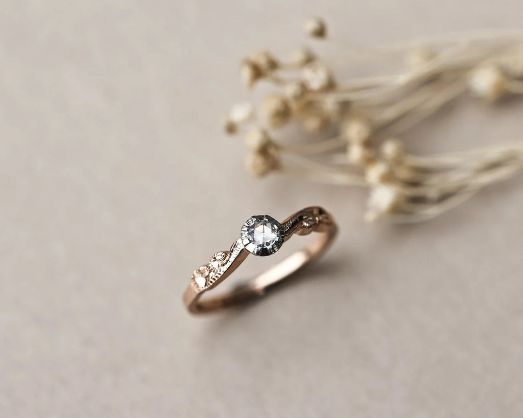 A delicate rose gold ring with a central clear gemstone and small side stones, styled on a neutral beige surface with dried flowers in the background.