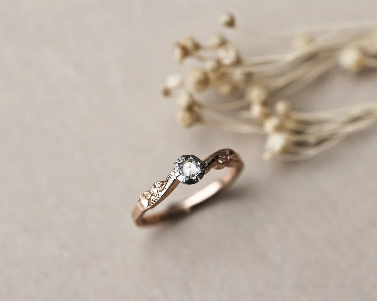 A delicate rose gold ring with a central clear gemstone and small side stones, styled on a neutral beige surface with dried flowers in the background.