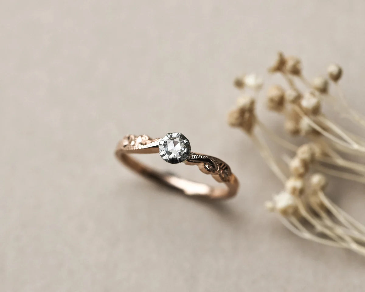 A delicate rose gold ring with an oval-cut clear gemstone and ornate filigree detailing, displayed on a neutral beige surface next to dried flowers.