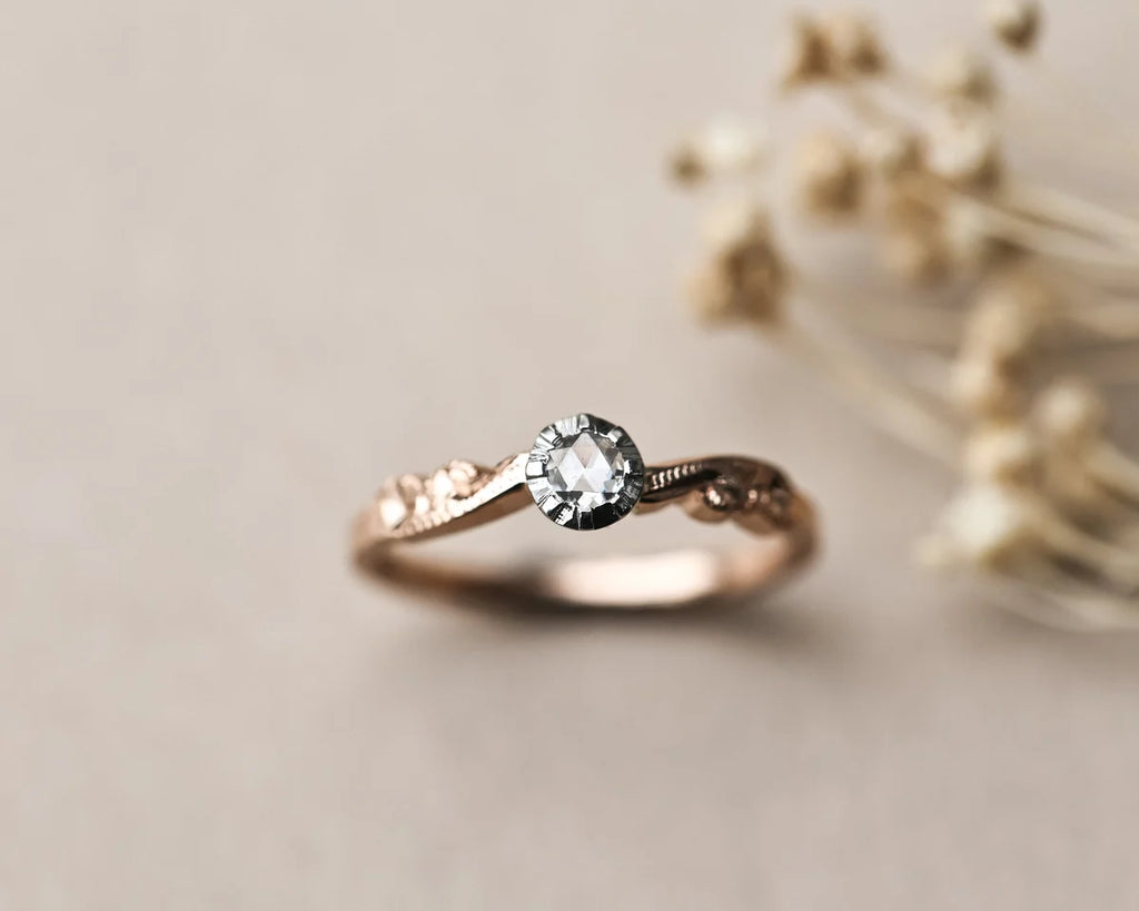 A delicate rose gold ring with a central clear gemstone and small side stones, resting on a neutral beige surface with blurred dried flowers in the background.