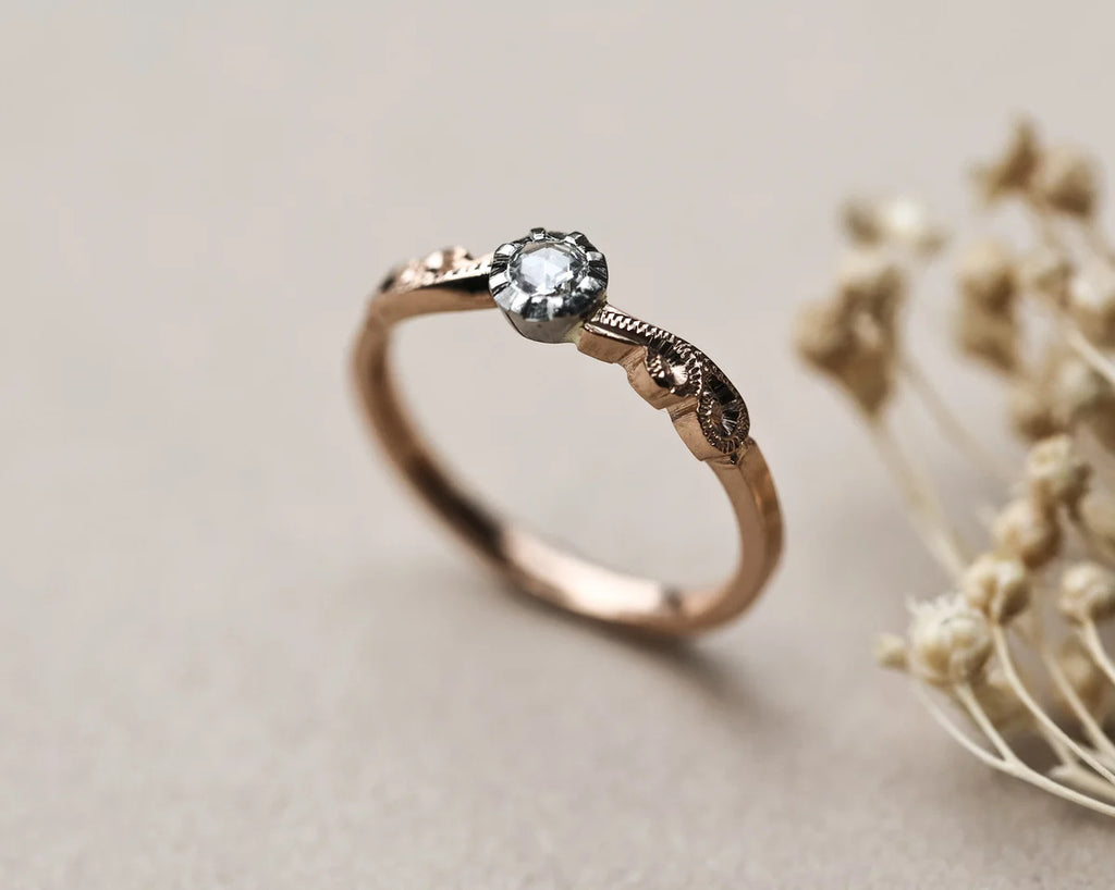 A rose gold solitaire ring with a clear round-cut gemstone and decorative engraved details on the band, shown on a neutral beige background with dried flowers as a prop.