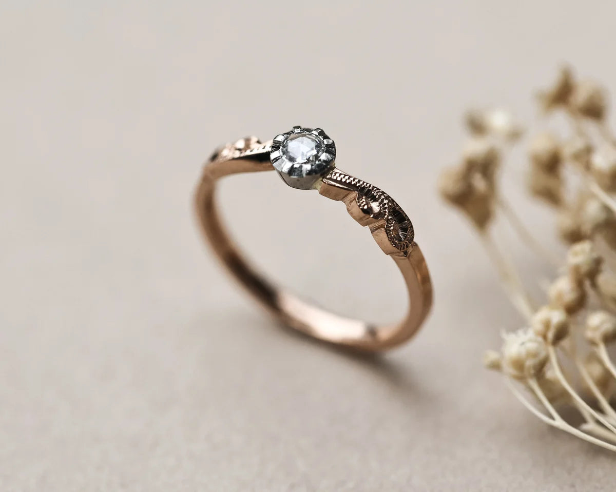 A rose gold solitaire ring with a clear round-cut gemstone and decorative engraved details on the band, shown on a neutral beige background with dried flowers as a prop.