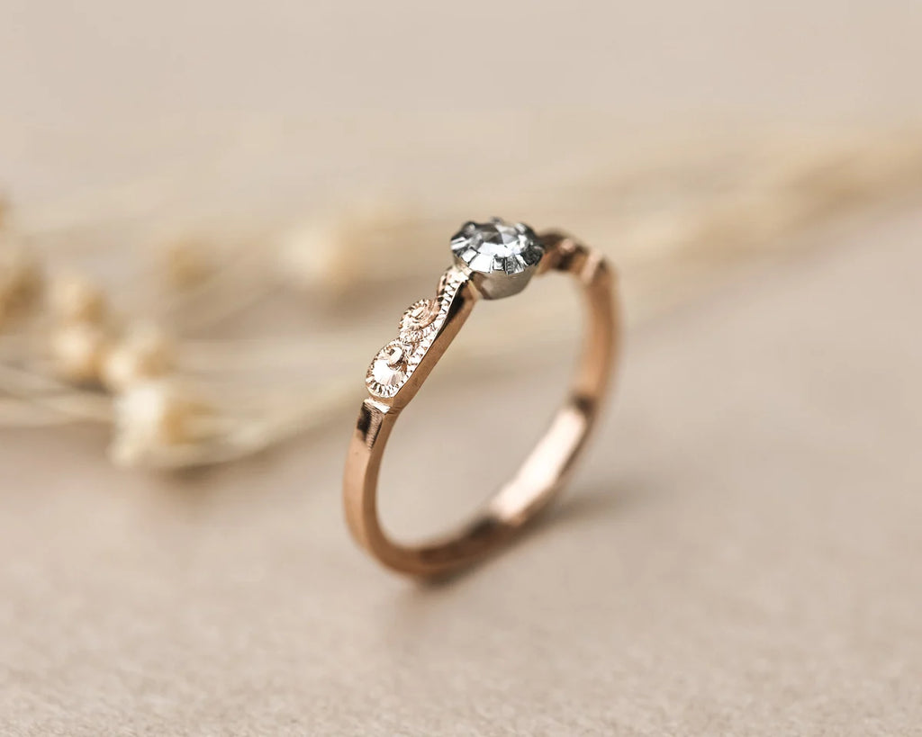 A rose gold solitaire engagement ring with a round clear gemstone, resting on a neutral beige surface with blurred dried flowers in the background.