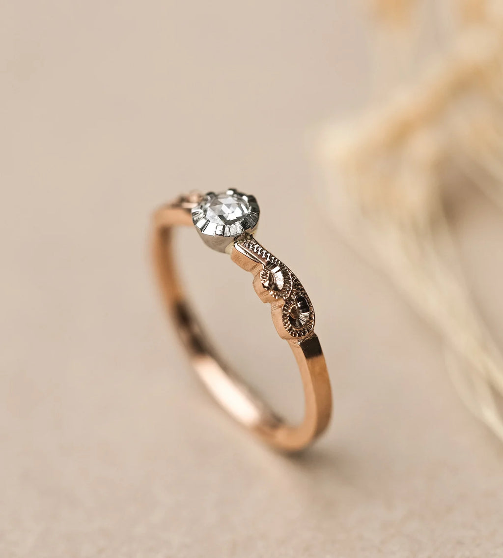 A delicate rose gold ring with a small clear gemstone and ornate filigree detailing, photographed on a soft beige background with blurred dried grass.
