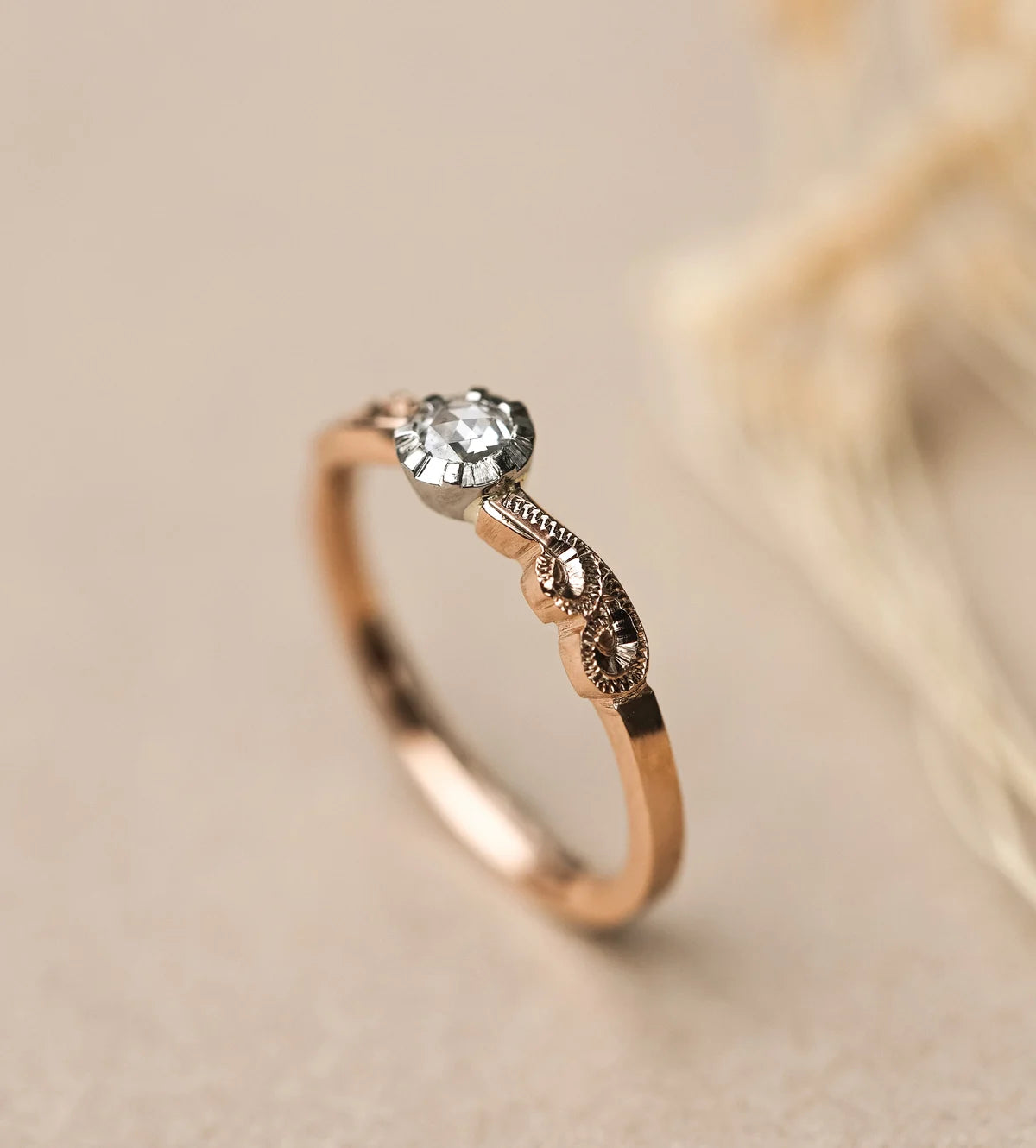 A delicate rose gold ring with a small clear gemstone and ornate filigree detailing, photographed on a soft beige background with blurred dried grass.