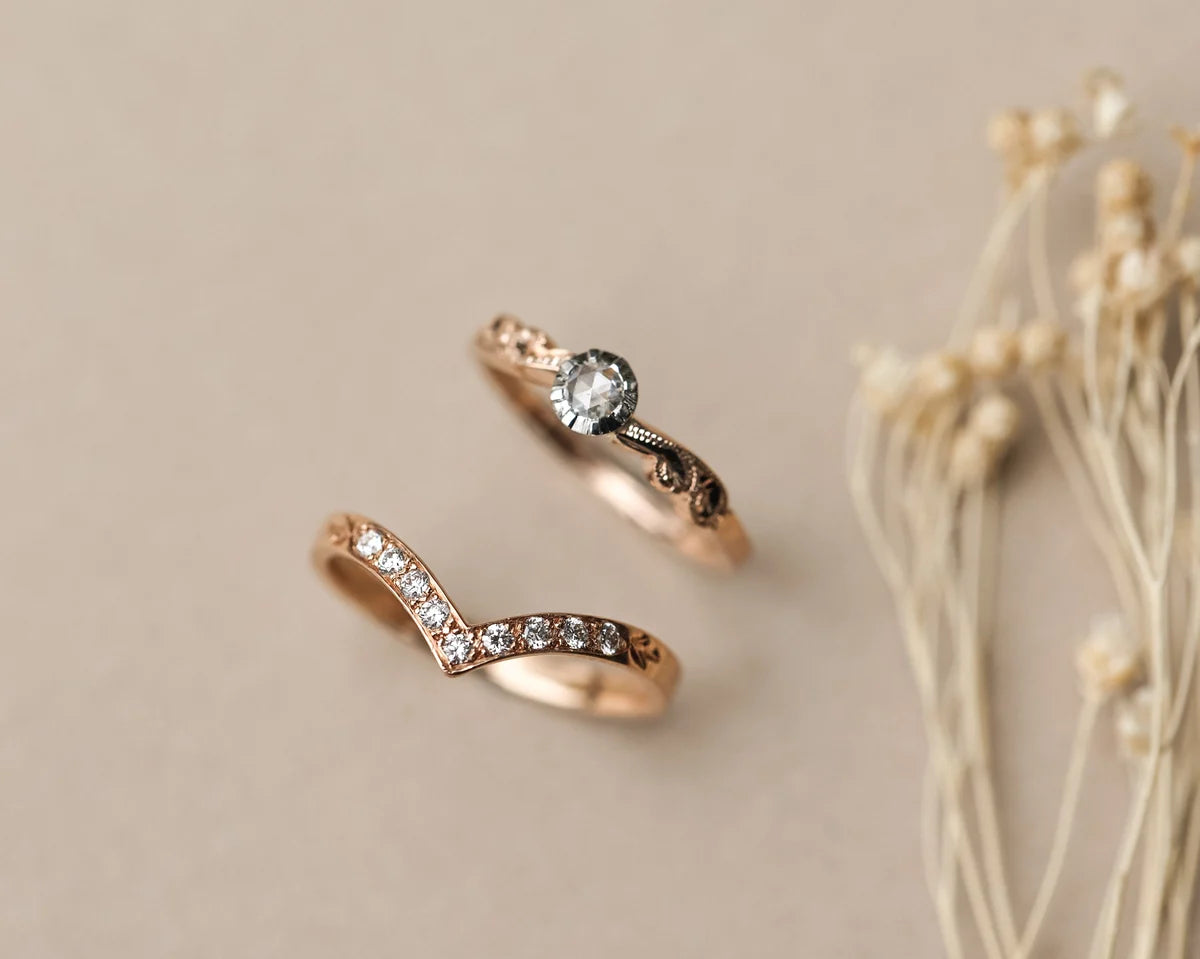 Two rose gold rings on a beige background, one with a single round clear gemstone and the other with a row of small clear stones along a wavy band, styled with dried flowers on the right.