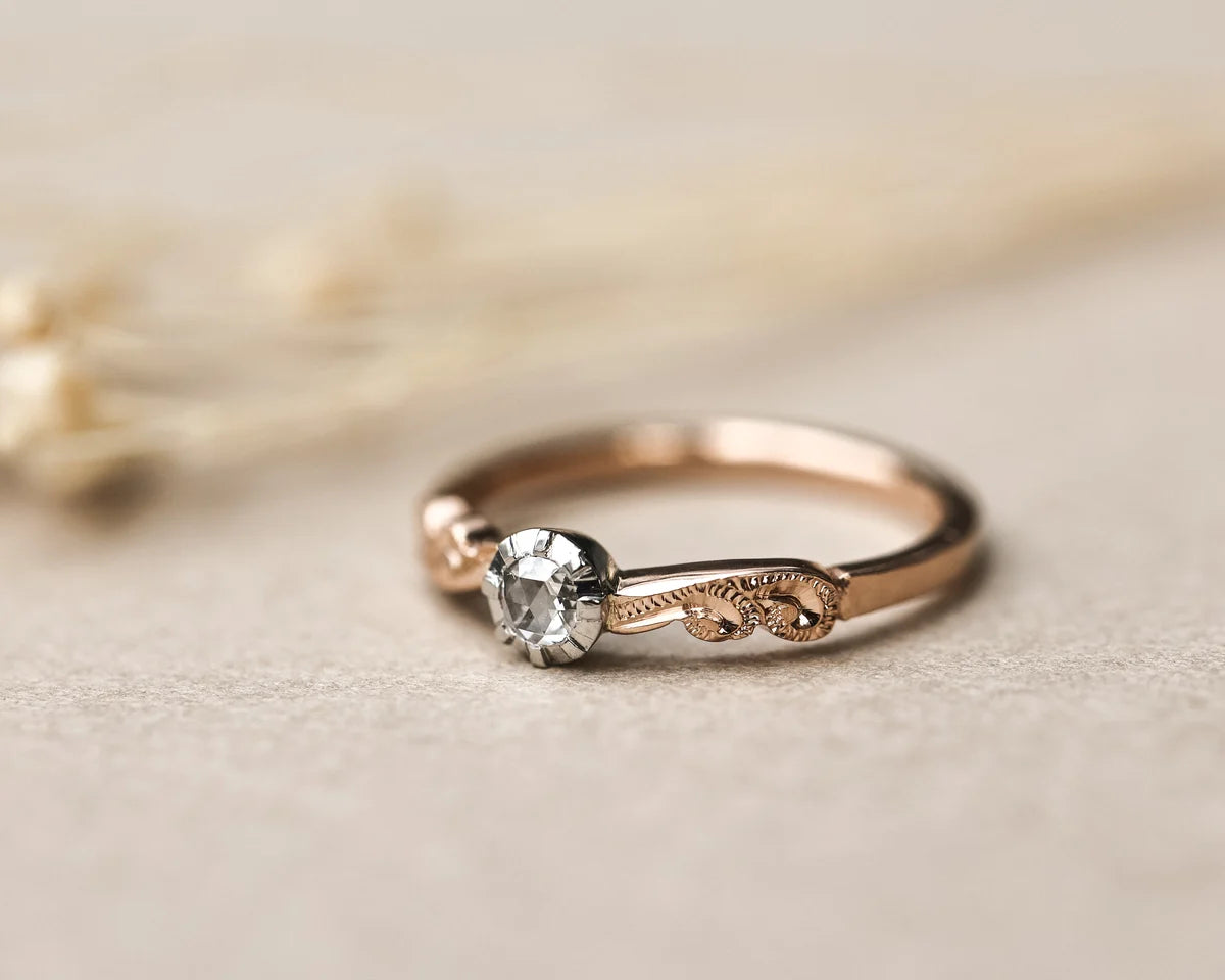 A delicate rose gold ring with a single clear gemstone and ornate scrollwork details on the band, resting on a textured beige surface with soft dried grass in the background.