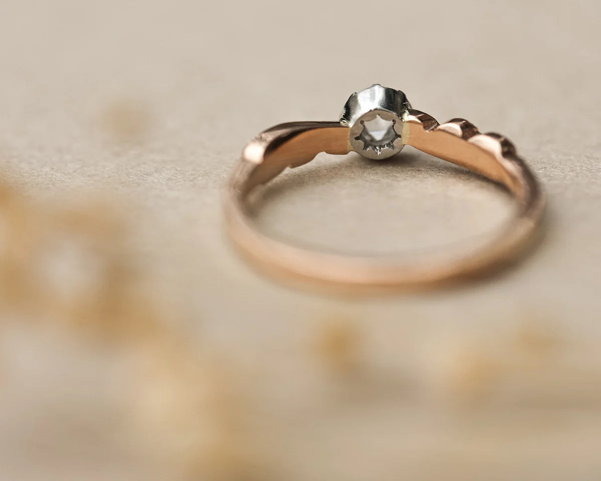 A delicate two-tone ring with a rose gold band and a silver-toned setting holding a small clear gemstone, resting on a soft beige textured surface.