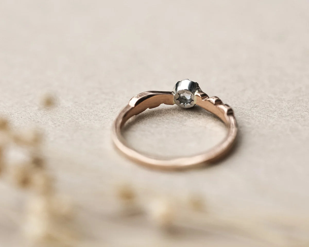 A delicate rose gold ring with a textured band and a small silver-toned bezel-set clear gemstone, resting on a neutral beige surface with soft blurred dried flowers in the foreground.