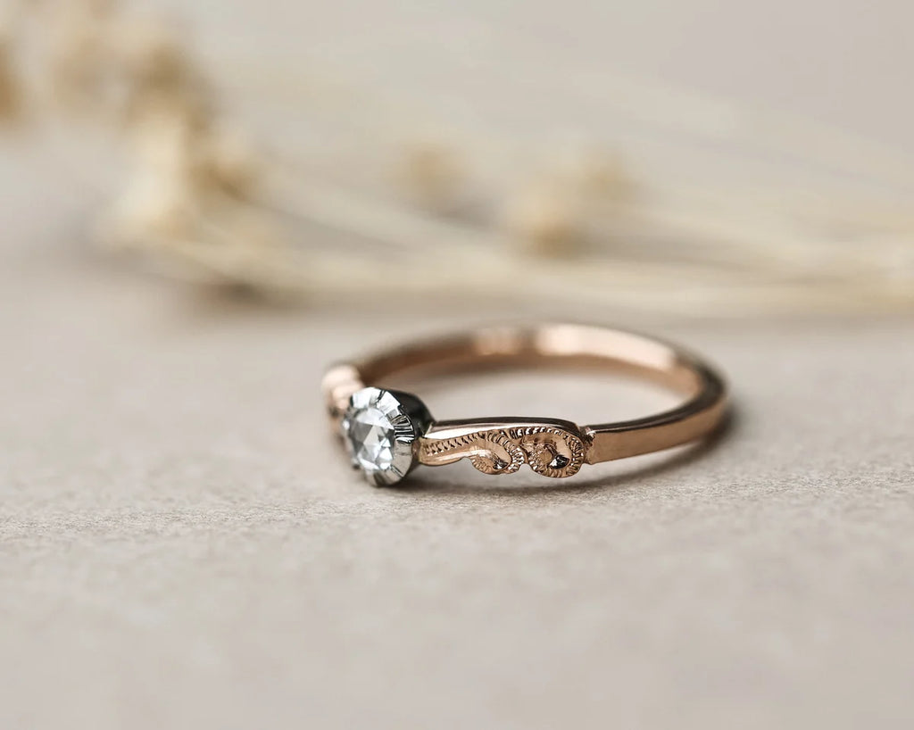 A delicate rose gold solitaire ring with an ornate engraved band and a clear round gemstone, resting on a textured beige surface with blurred dried flowers in the background.