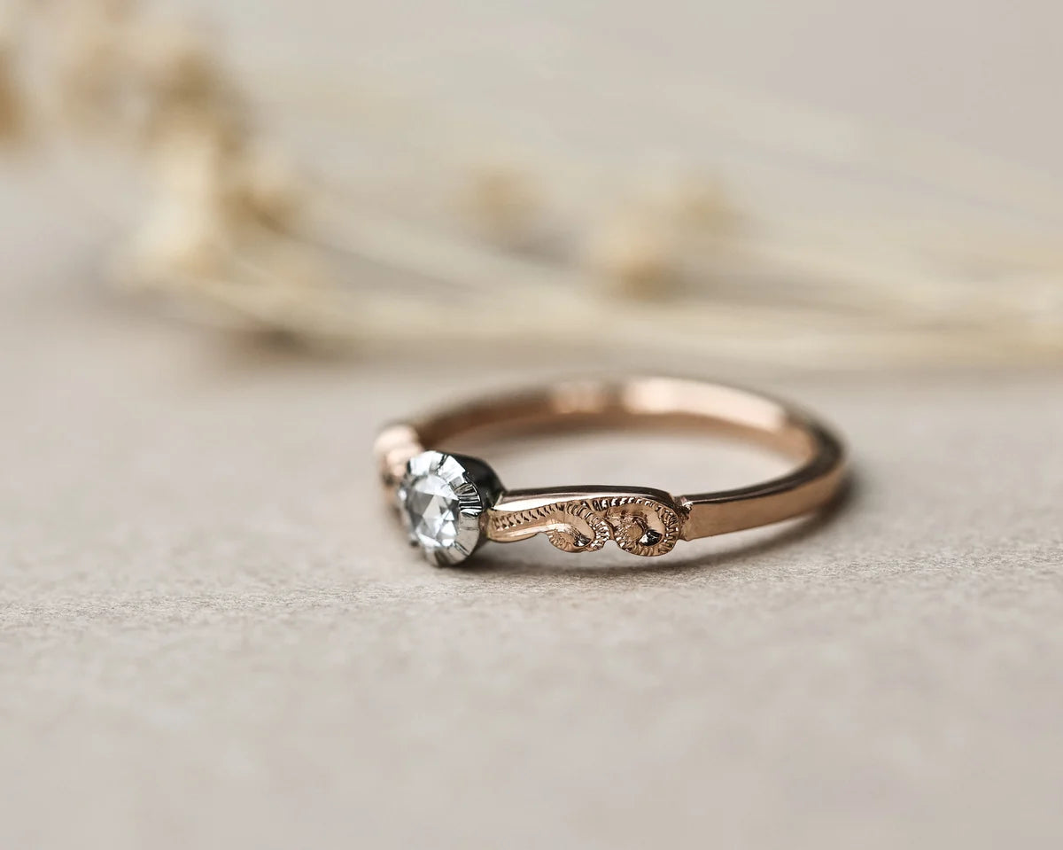 A delicate rose gold solitaire ring with an ornate engraved band and a clear round gemstone, resting on a textured beige surface with blurred dried flowers in the background.