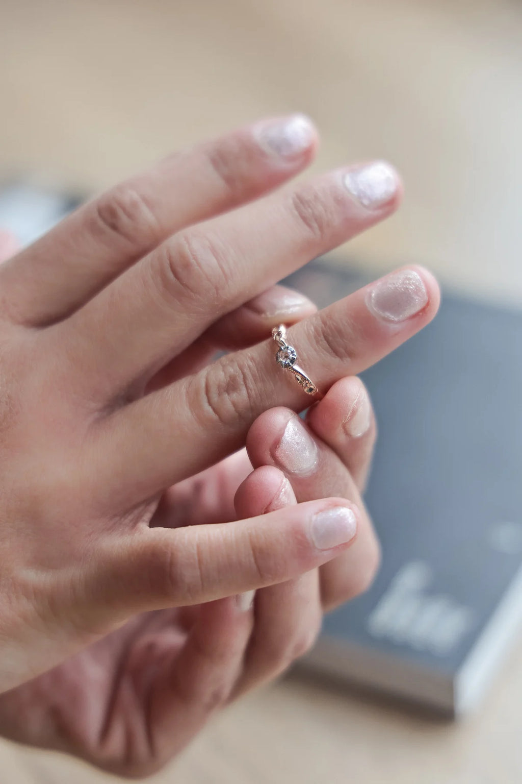 A close-up of a person's hands holding a delicate silver-toned ring with a small clear gemstone, set against a softly blurred background.