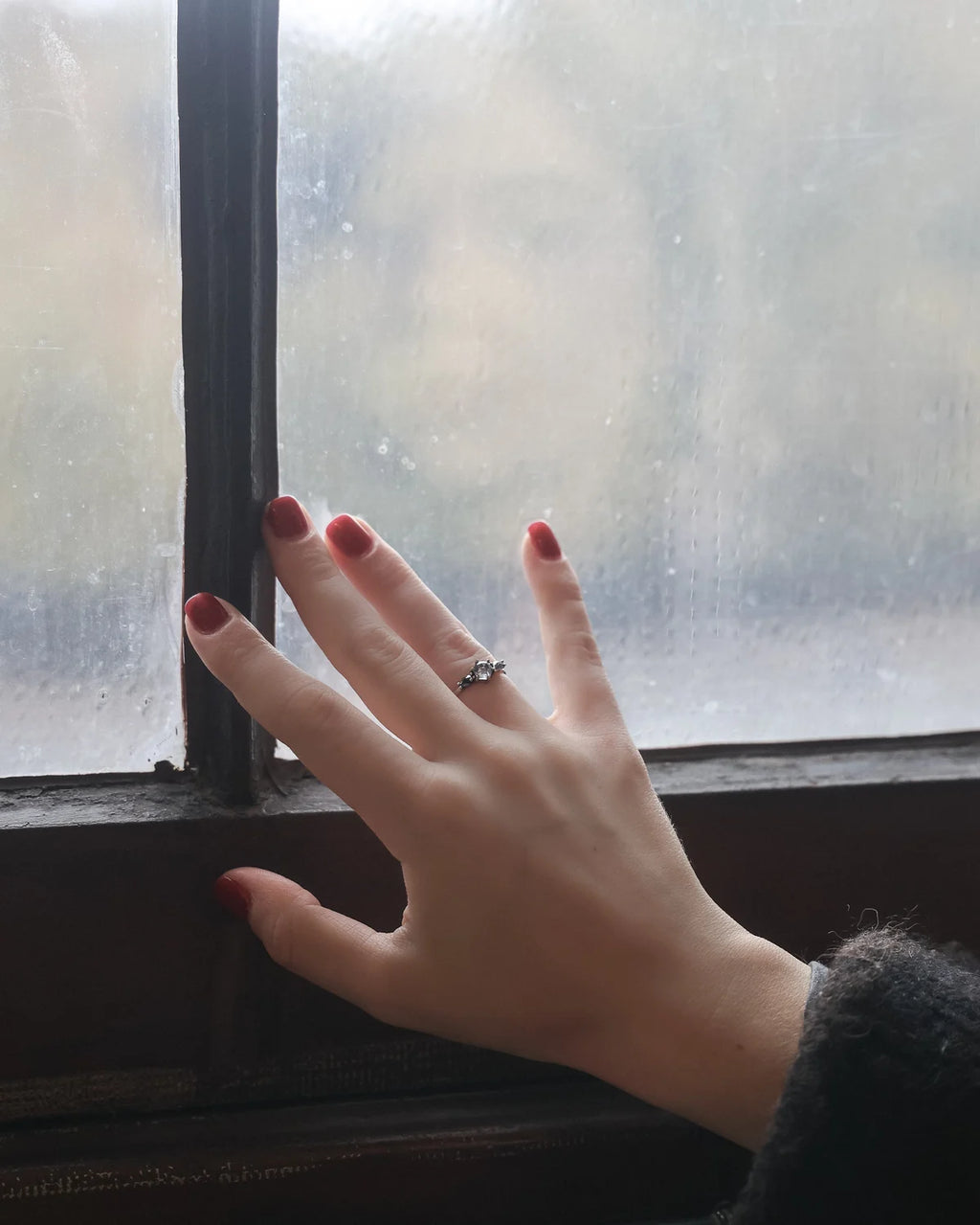 A close-up of a person's hand resting on a window frame, wearing a silver ring with small clear stones. The hand has dark red painted nails, and the background is a soft-focus, misty window view.
