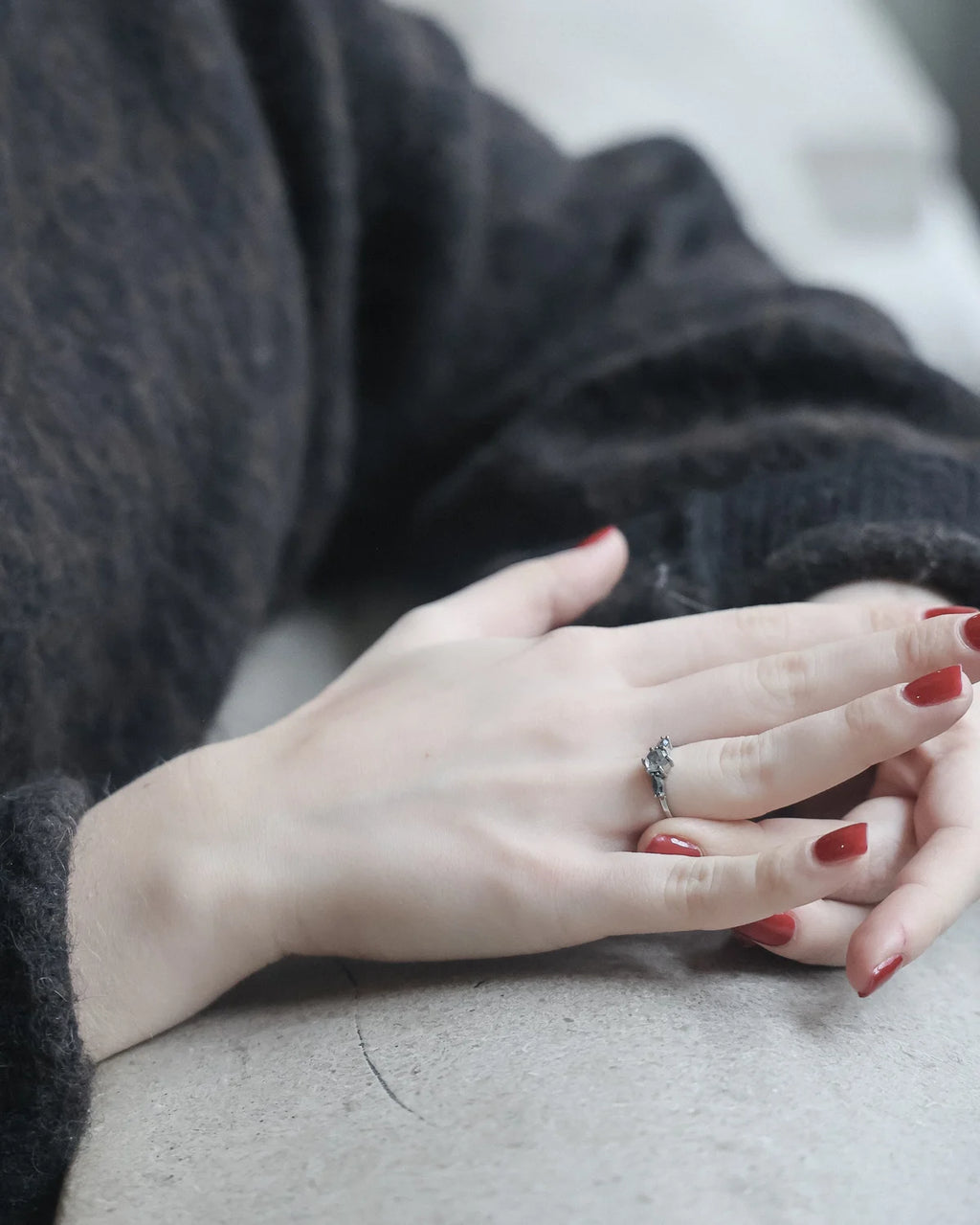 A close-up of a person's hands resting on a light gray surface, wearing a silver ring with a small clear gemstone. The person is wearing a dark charcoal gray knit sweater, and their fingernails are painted red.