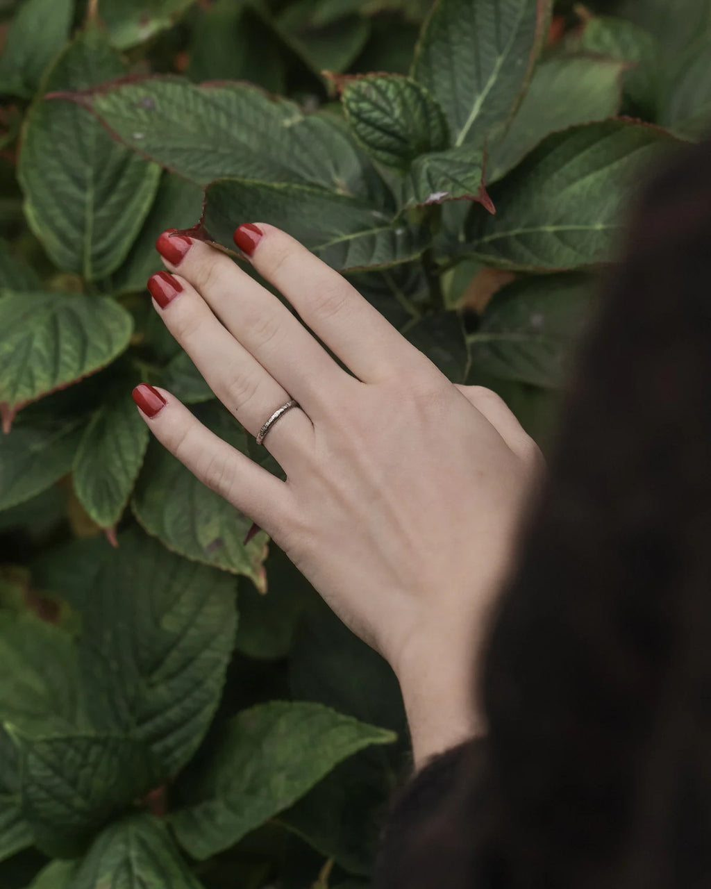 Hand with red nail polish and a ring on a leafy background