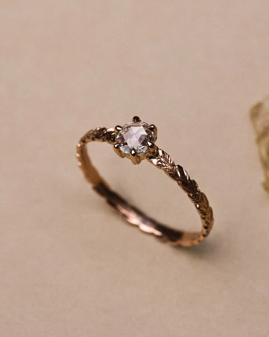 A delicate rose gold ring with a textured, twisted band and a central clear gemstone, displayed on a neutral beige background.