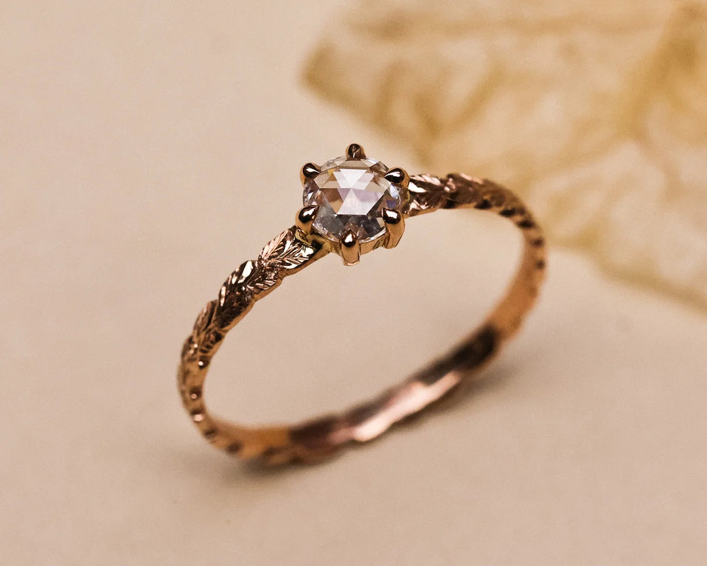 A close-up of a rose gold ring with a round-cut clear gemstone in a prong setting, resting on a beige surface with a soft-focus decorative background.