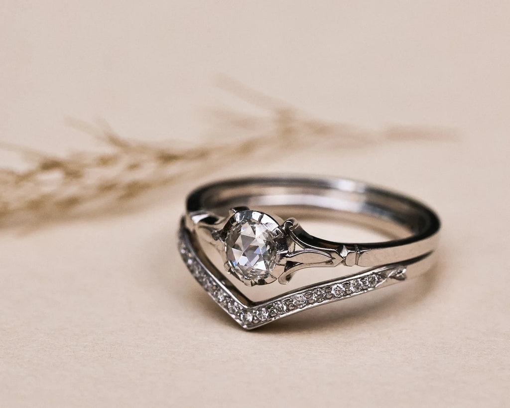 A silver-toned ring with a round-cut clear center stone and a curved pavé band, photographed on a warm beige surface with soft blurred dried stems in the background.