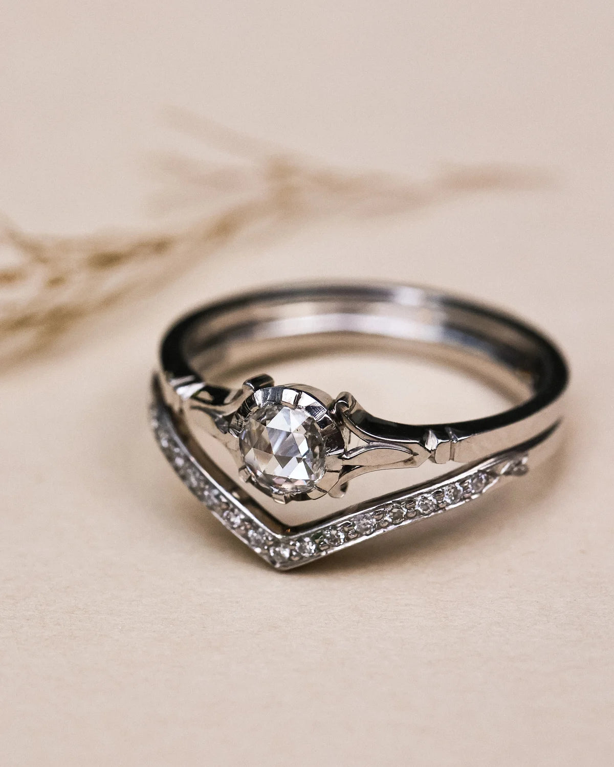 A silver-toned ring with a large oval clear gemstone in a prong setting, accented by pavé stones along a V-shaped band, photographed on a warm beige background.