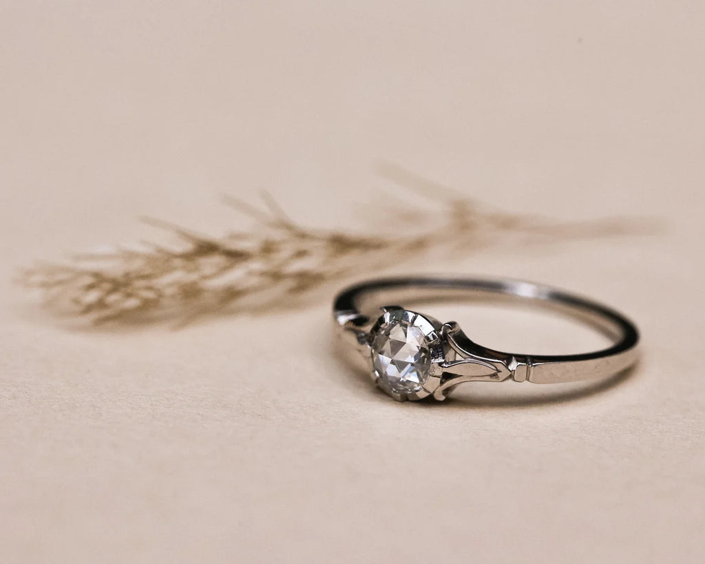 A silver ring with a clear faceted gemstone in a prong setting, resting on a textured beige surface with dried wheat stalks in the background.