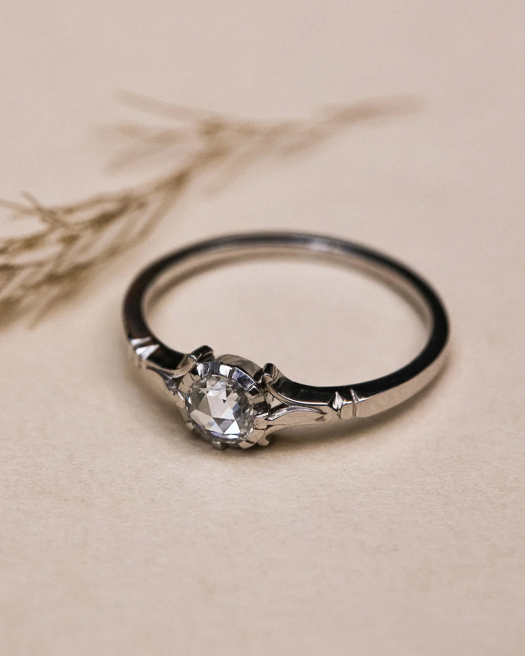 A silver solitaire ring with a clear faceted gemstone, resting on a textured beige surface with dried grass in the background.