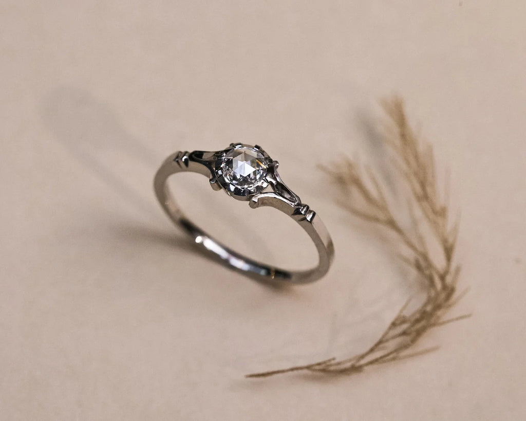 A silver ring with a clear round-cut gemstone in a decorative setting, resting on a beige surface next to dried grass.