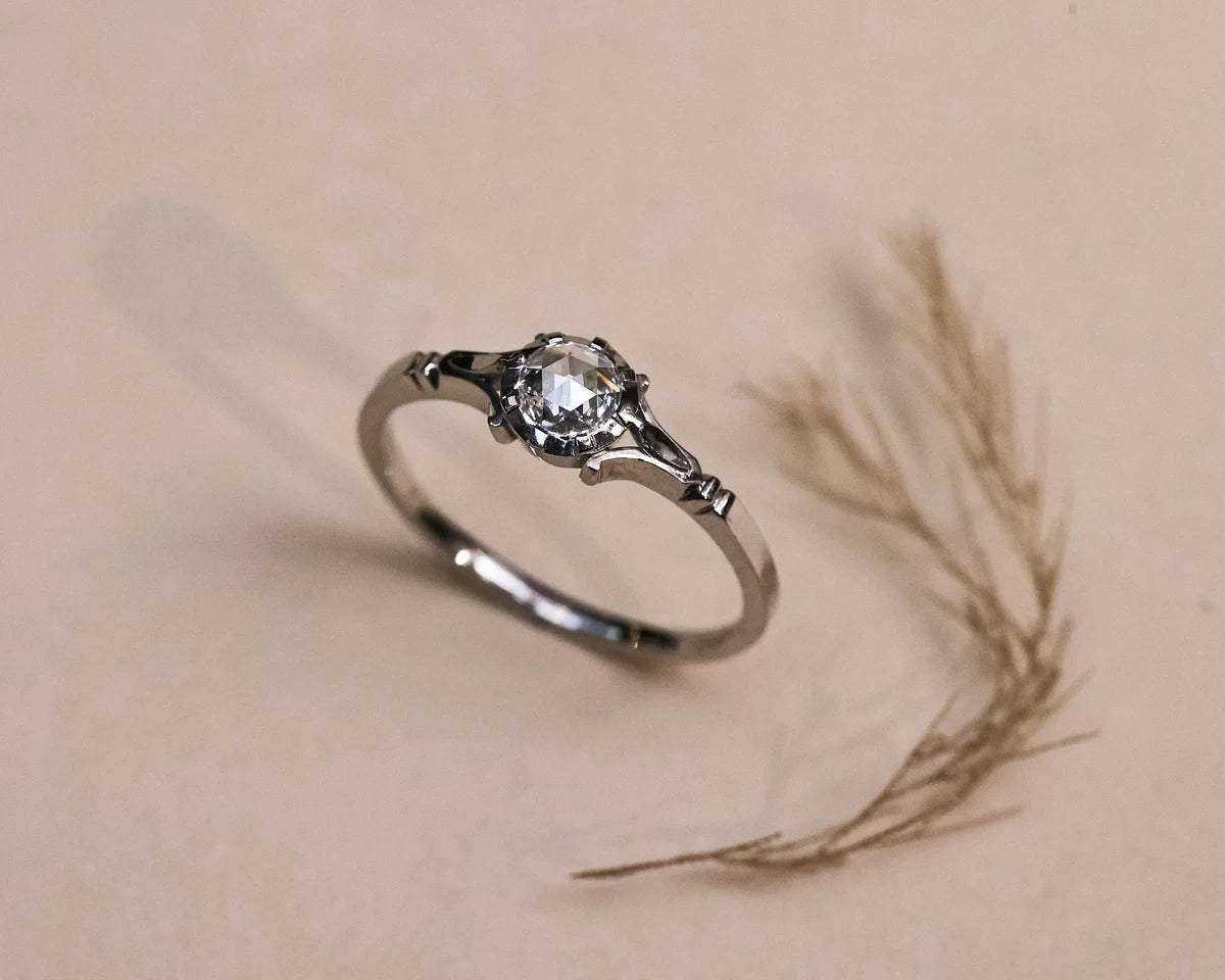 A silver ring with a clear round-cut gemstone in a decorative setting, resting on a beige surface next to dried grass.