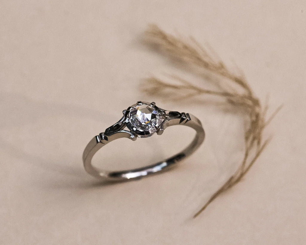 A silver-toned ring with a clear round gemstone in an ornate setting, photographed on a warm beige surface with dried grass stems behind it.