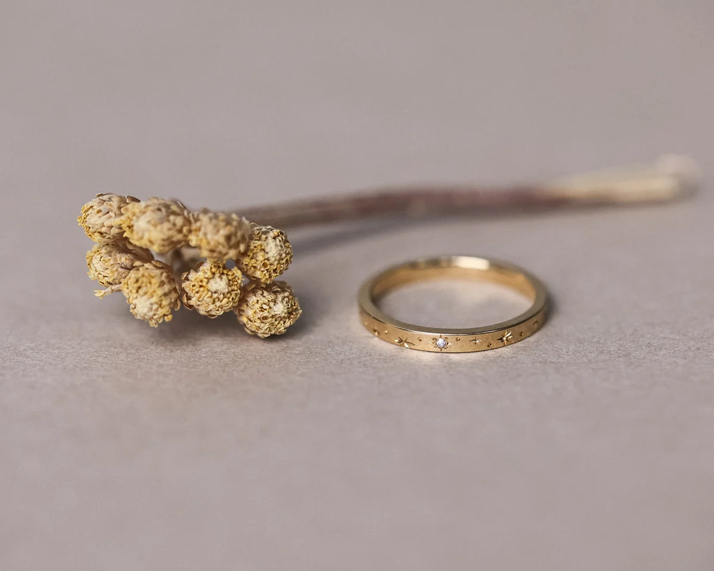 Gold ring with engraved details next to a dried flower on a neutral background