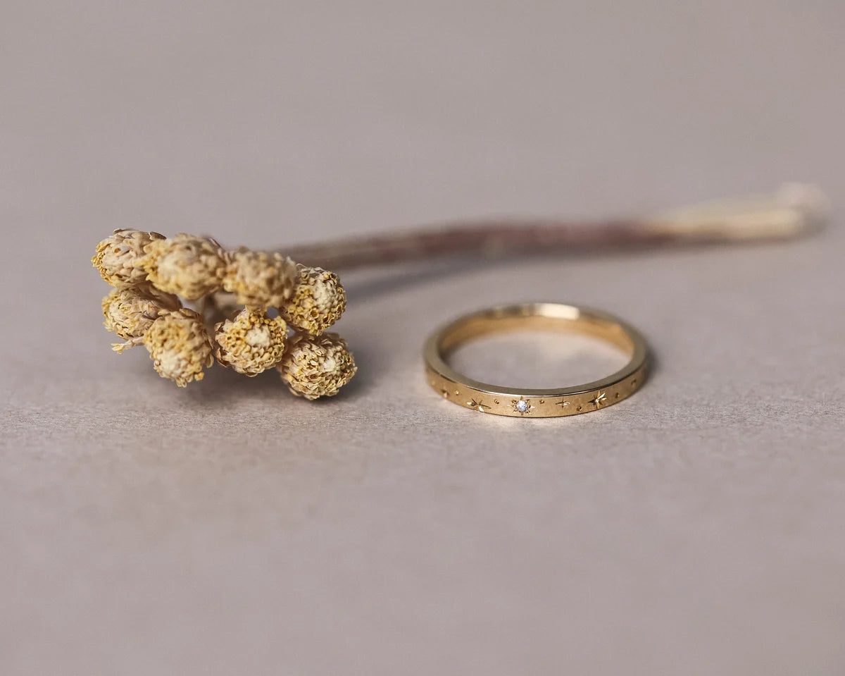 Gold ring with engraved details next to a dried flower on a neutral background