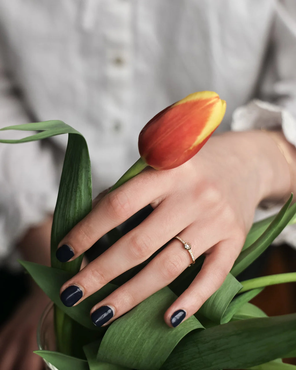 A close-up of a hand with dark navy blue nail polish resting on green tulip leaves, wearing a delicate gold ring with a small clear gemstone. A red and yellow tulip bud is held near the hand, and a person in a white button-up shirt is softly blurred in the background.