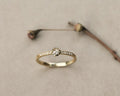 A delicate yellow gold ring with a central round-cut diamond and small pavé diamonds along the band, resting on a textured beige surface with a blurred dried flower stem in the background.
