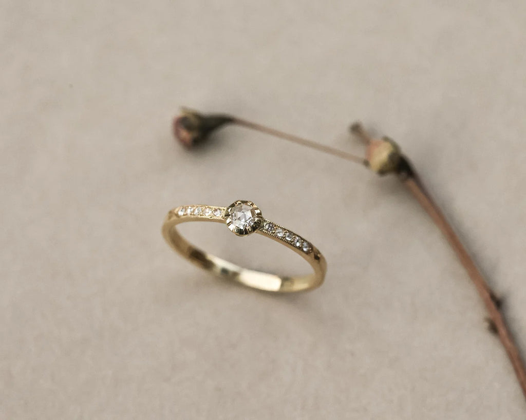 A delicate yellow gold ring with a small round-cut center stone and tiny pavé-set side stones, resting on a neutral beige textured surface with dried botanical stems in the background.