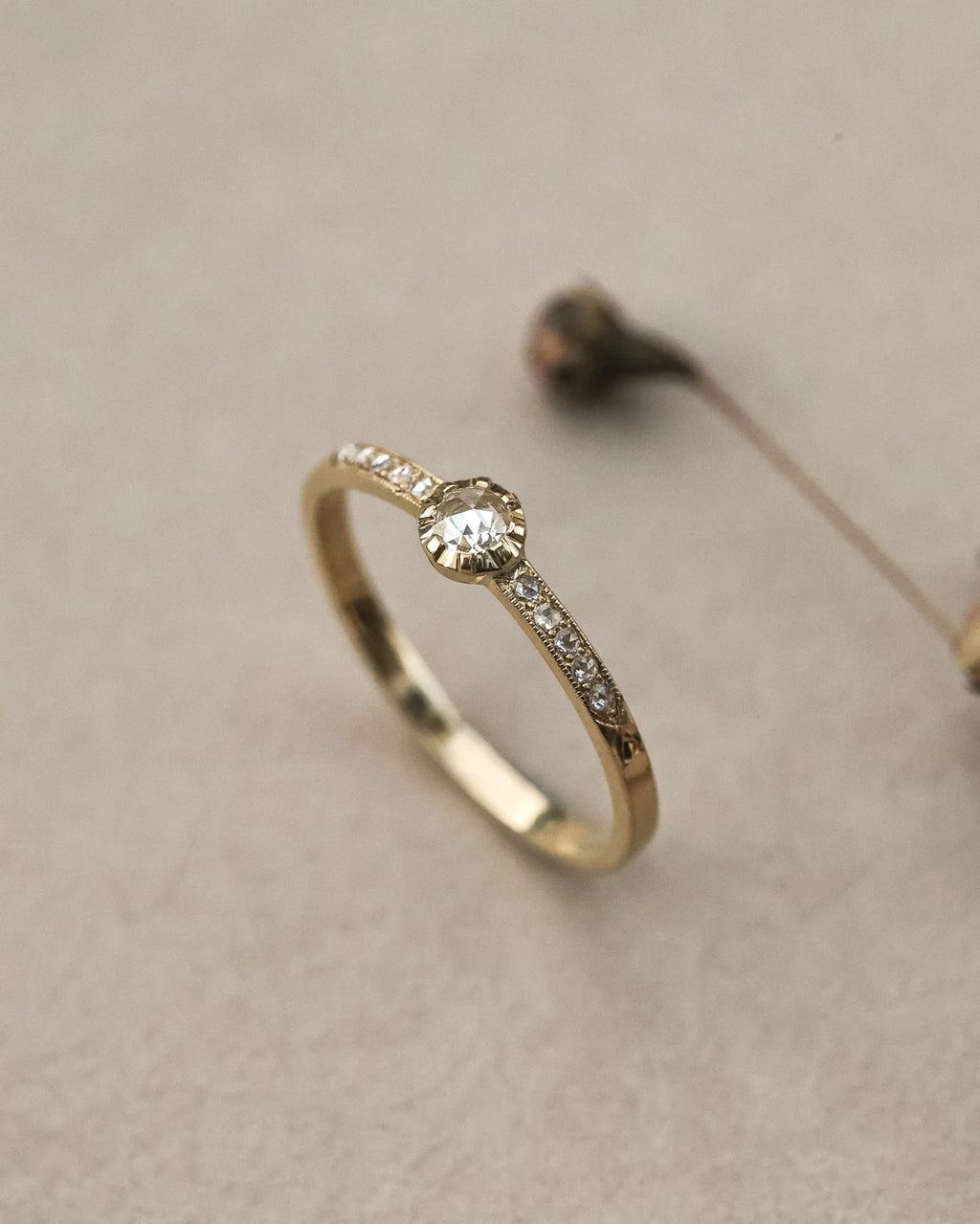 A gold ring with a round-cut clear center stone and a pavé band of small clear stones, photographed on a soft beige textured surface.