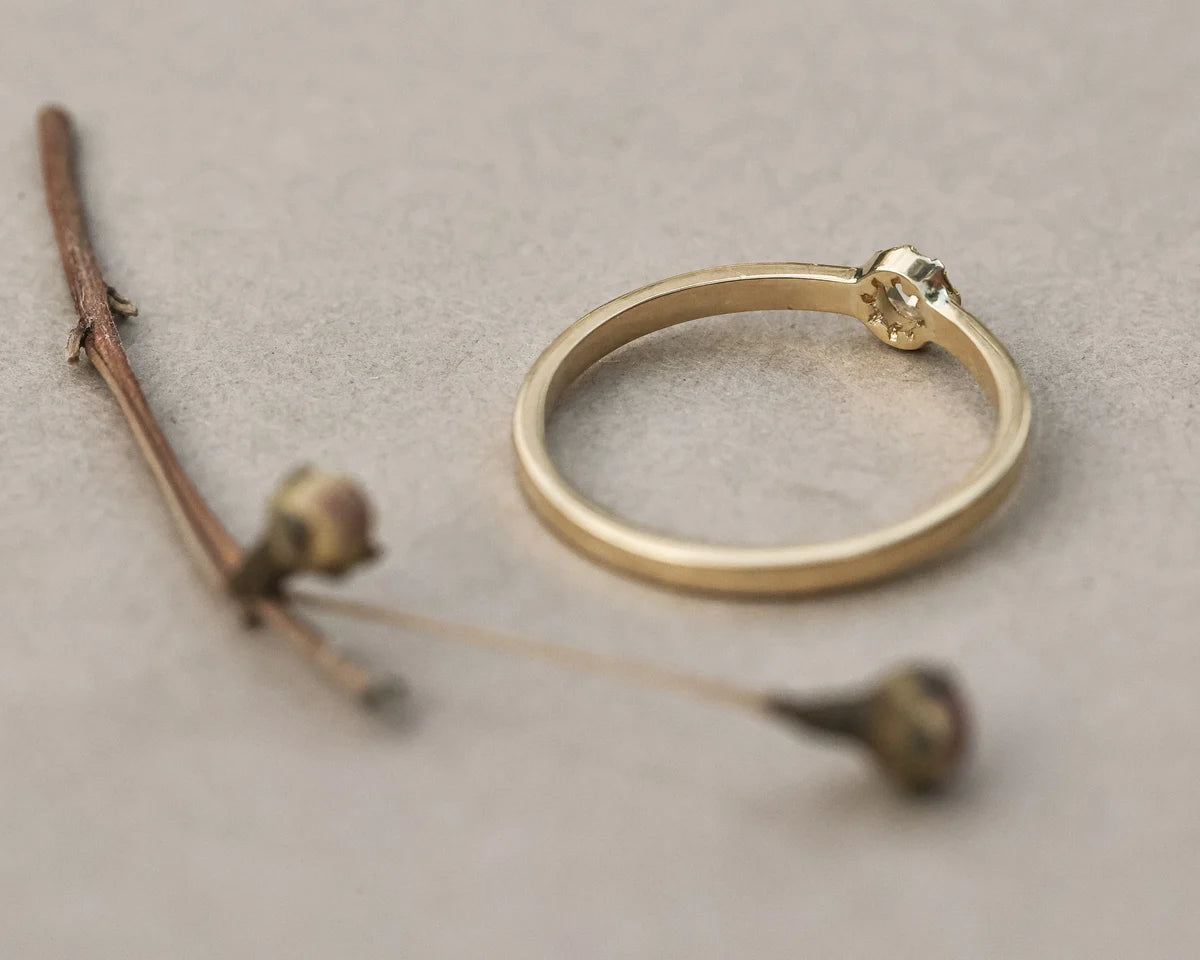 A minimalist gold ring with a small textured gold accent at the top, displayed on a neutral gray textured surface next to dried botanical stems.