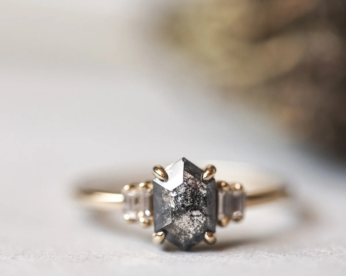 A close-up of a gold ring featuring a central dark gray to black textured gemstone, flanked by two small clear baguette side stones, set against a soft, blurred light background.