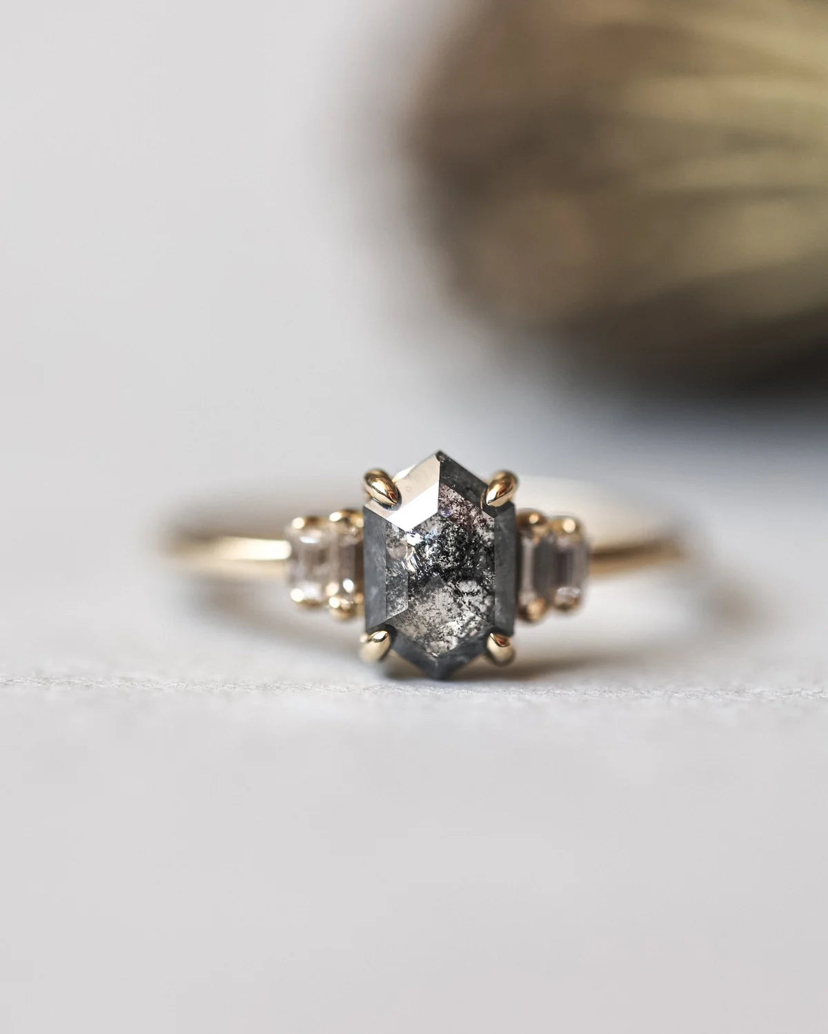 A close-up shot of a gold ring featuring a central dark gray, sparkly gemstone flanked by two small clear stones on each side, set against a soft-focus neutral background.