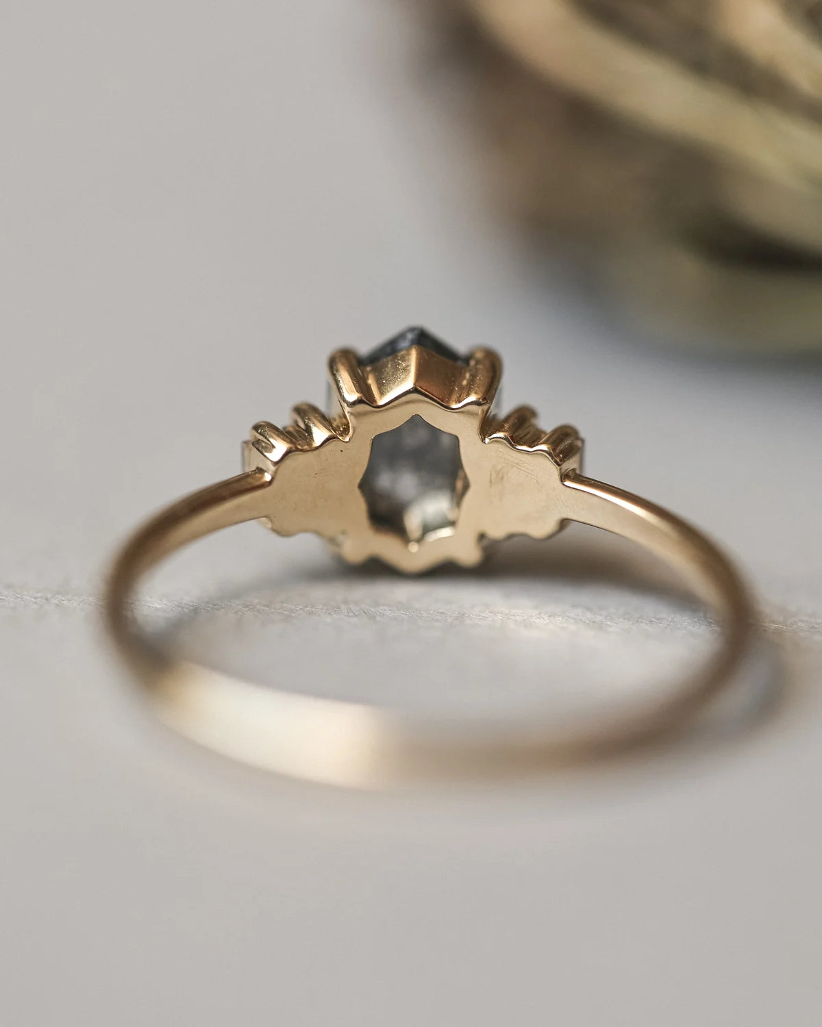 A gold ring with a unique, sculptural crown-like setting holding a small dark gray gemstone, photographed on a soft light gray surface.