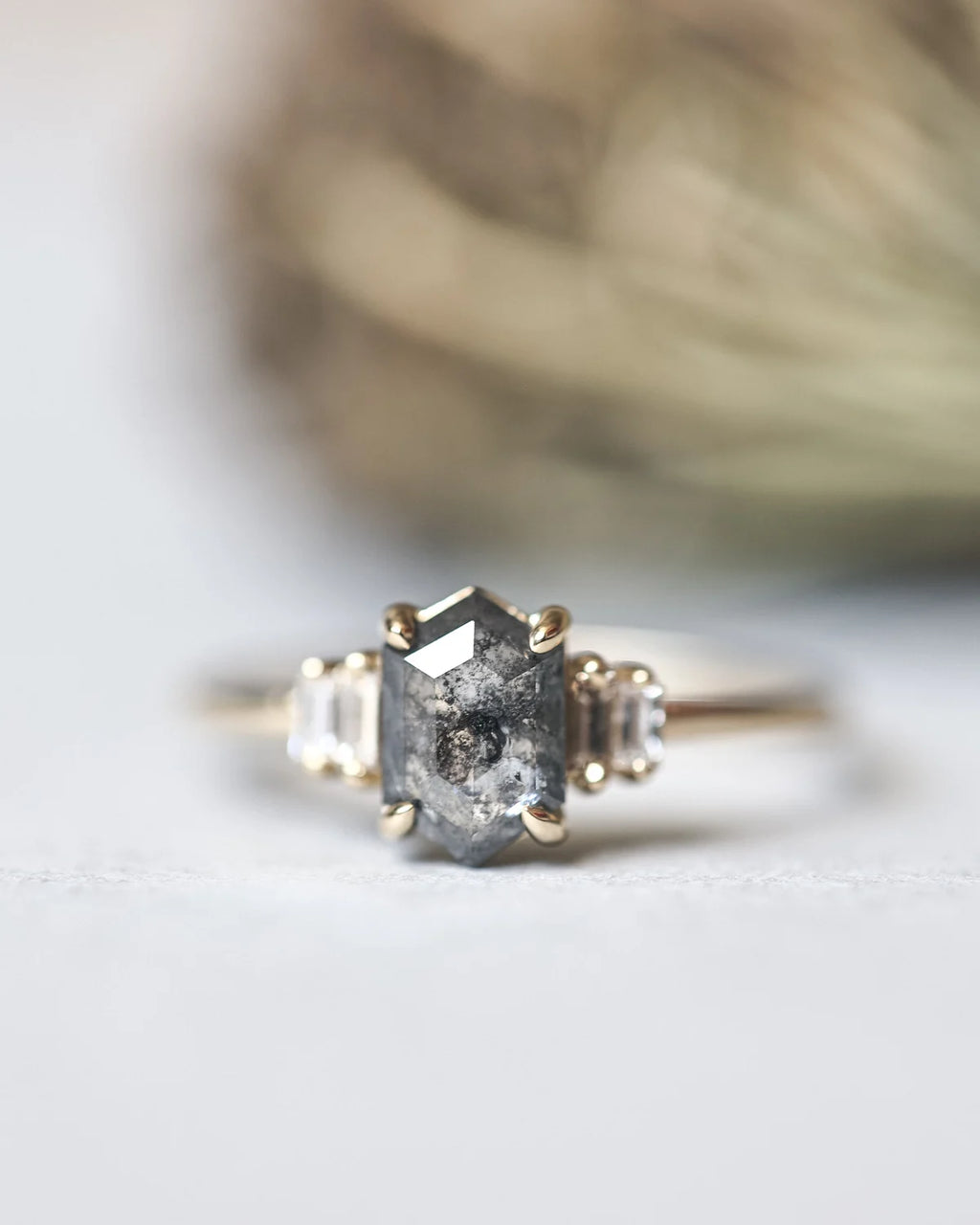 A close-up shot of a gold ring featuring a central gray baguette-cut gemstone with inclusions, flanked by two small clear round-cut side stones, set against a soft-focus neutral background.