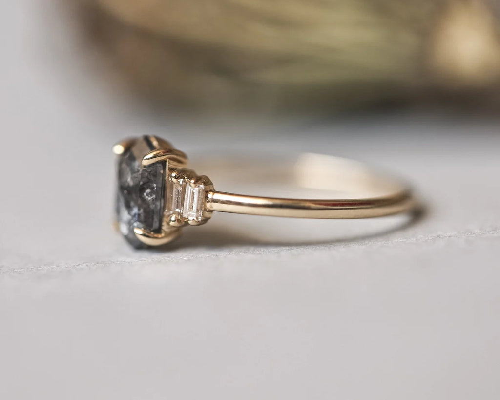 A close-up shot of a gold ring featuring a large, rough-cut dark gray gemstone with baguette side stones, resting on a light gray surface.