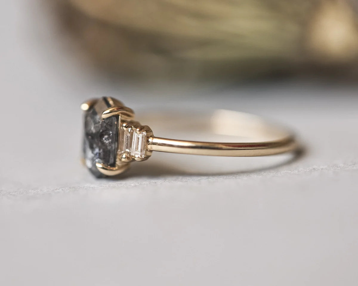 A close-up shot of a gold ring featuring a large, rough-cut dark gray gemstone with baguette side stones, resting on a light gray surface.