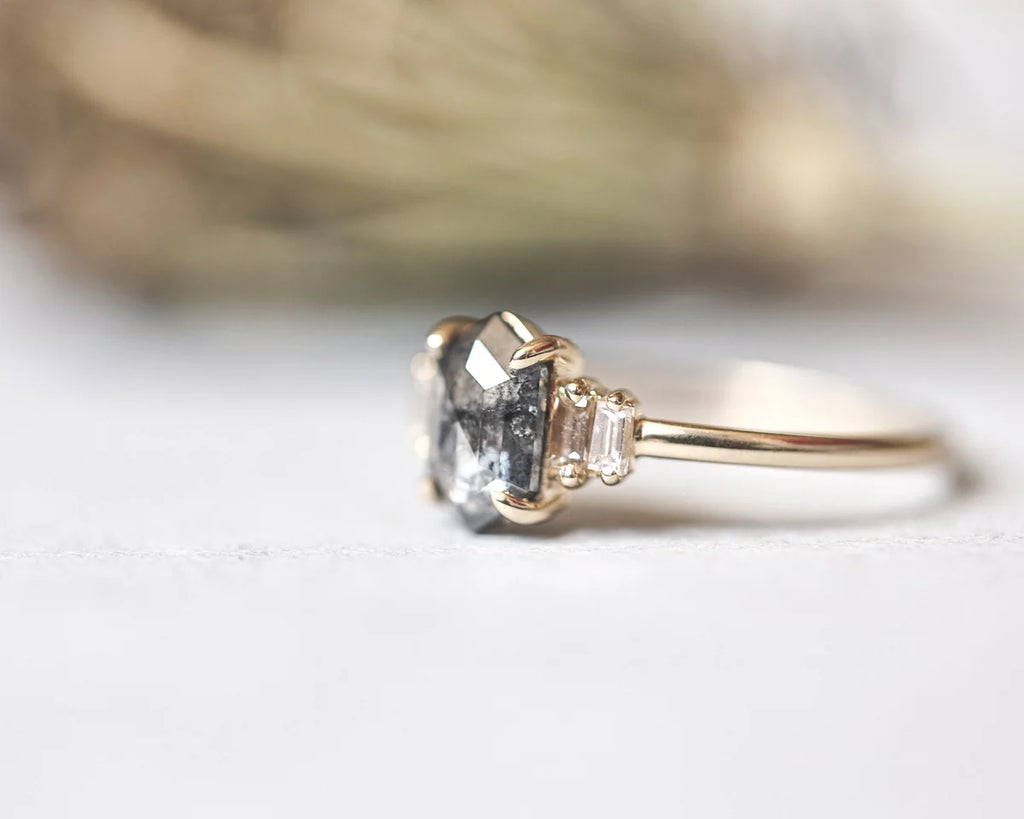 A close-up shot of a gold ring featuring a large clear faceted center stone and two small clear accent stones on either side of the band, set against a soft, out-of-focus neutral background.