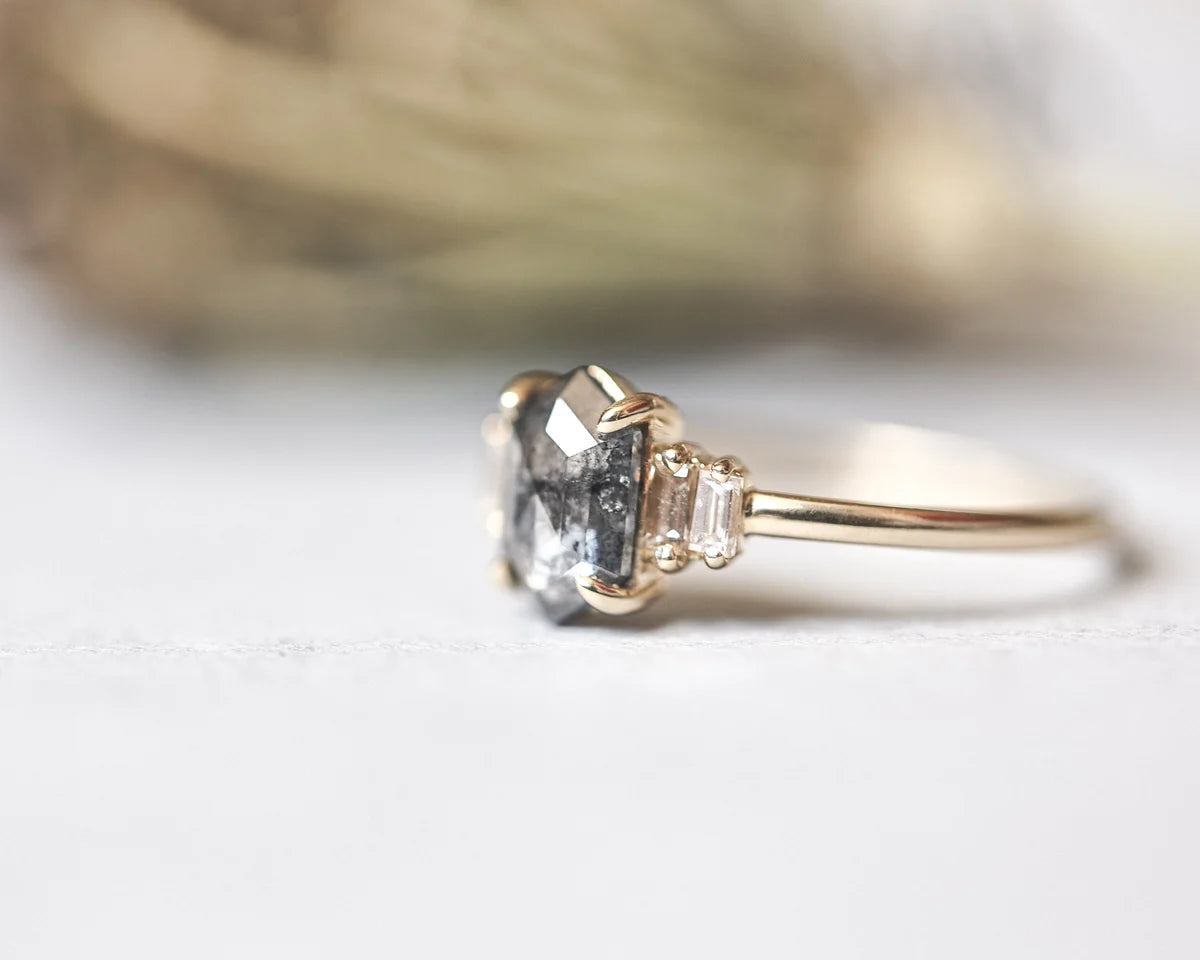 A close-up shot of a gold ring featuring a large clear faceted center stone and two small clear accent stones on either side of the band, set against a soft, out-of-focus neutral background.