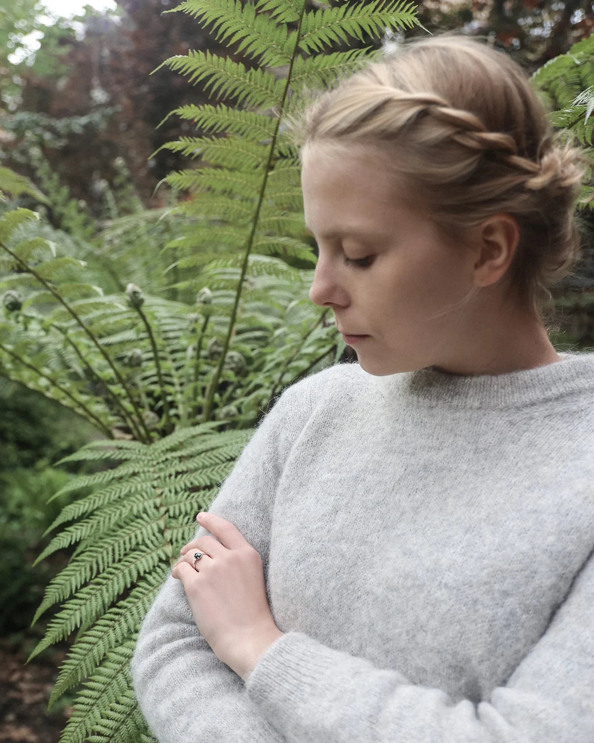 A woman with blonde hair in a braided bun wearing a light gray knit sweater, standing outdoors in front of green ferns.