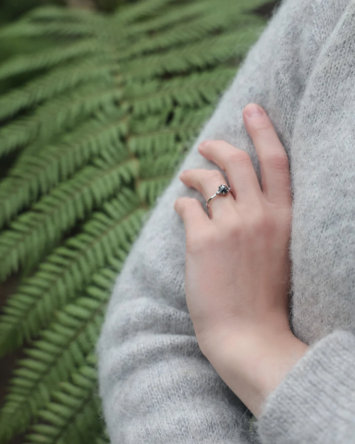 A close-up of a person's hand resting on their gray knit sweater, wearing a silver-toned ring with a small dark gemstone, set against a blurred green fern background.