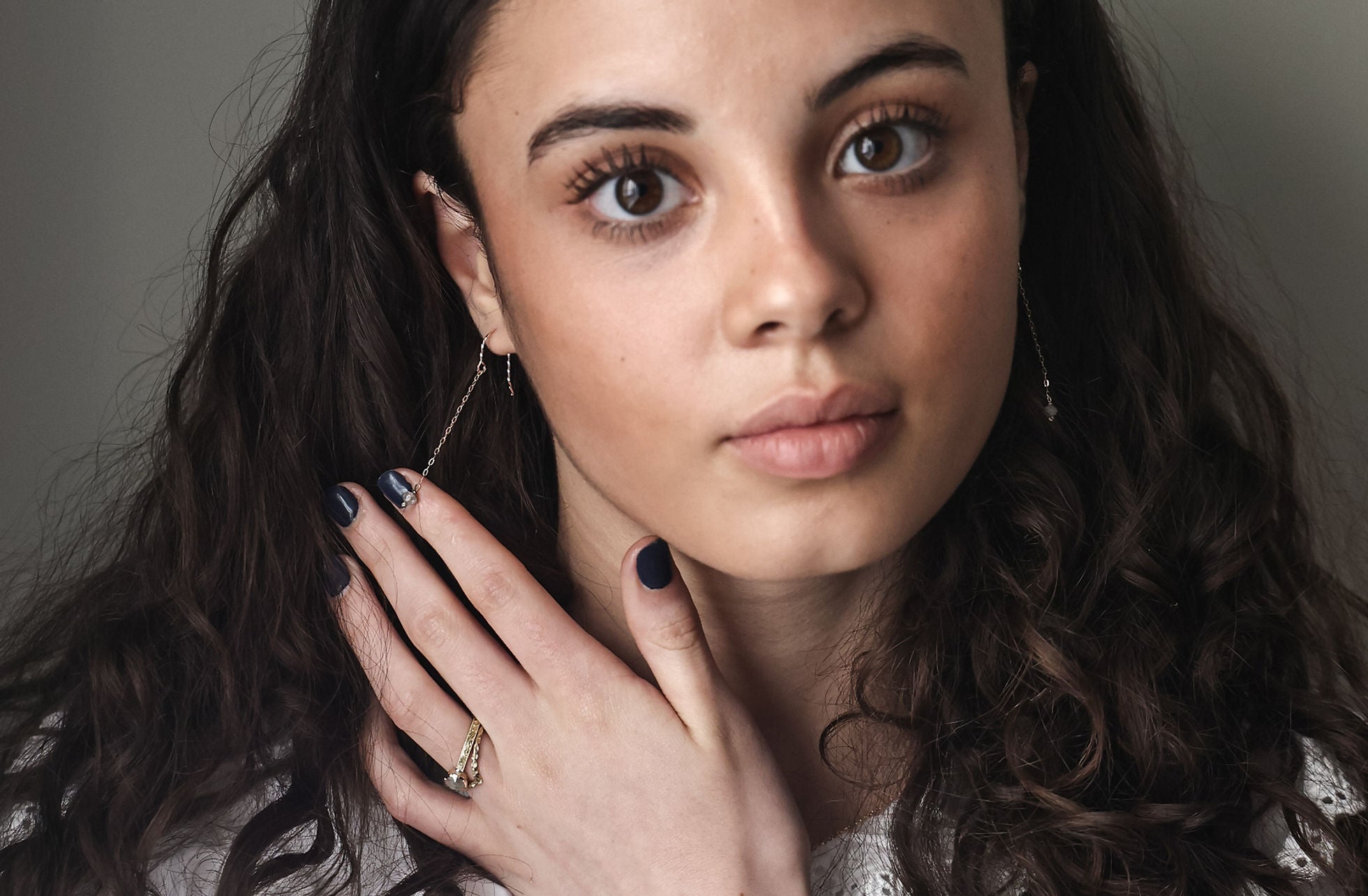 Woman with dark hair and jewelry, including earrings and a ring, against a neutral background.